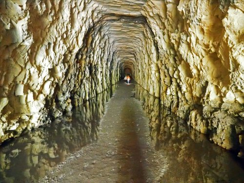 A long, narrow tunnel with textured stone walls and water reflecting the ceiling, illuminated by soft light.