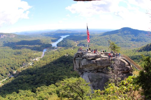 A scenic view from a rocky overlook with an American flag, surrounded by lush green mountains and a winding river below.