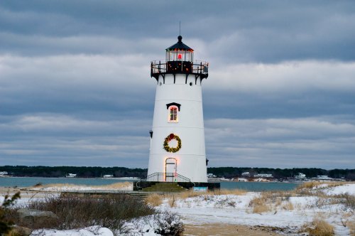 A white lighthouse adorned with a wreath, standing by the water under a cloudy sky, surrounded by snow and grass.