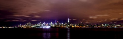A panoramic view of a city skyline at night, illuminated buildings reflecting on the water under a cloudy sky.