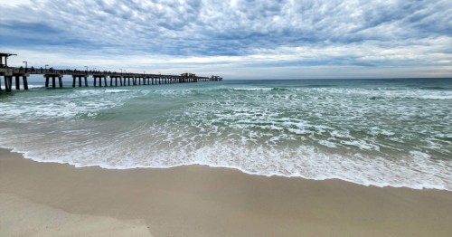 A serene beach scene with gentle waves, a cloudy sky, and a pier extending into the ocean.