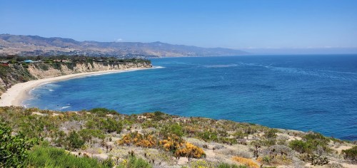 A scenic coastal view featuring a sandy beach, clear blue water, and rolling hills under a bright blue sky.