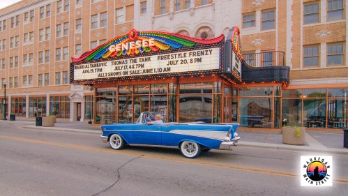 A classic blue car drives past the historic Genesee Theatre marquee advertising upcoming shows.
