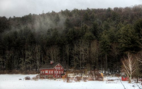 A red barn stands in a snowy field, surrounded by misty trees and a forested hillside in the background.