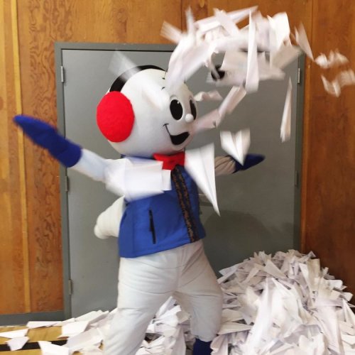 A cheerful mascot in a blue outfit throws paper confetti in the air, surrounded by a pile of shredded paper.