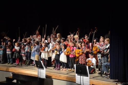 A group of children performing on stage with various instruments, including violins and guitars, during a music event.