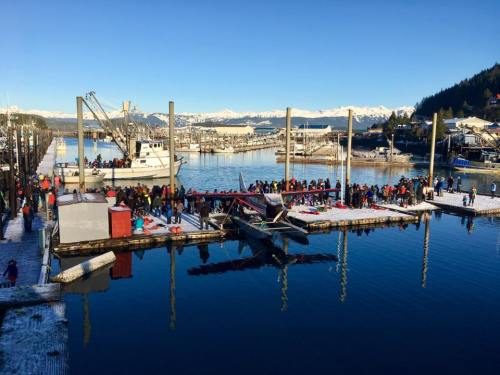 A busy harbor scene with a seaplane docked, surrounded by people and snow-capped mountains in the background.