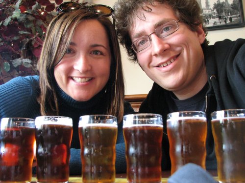 A woman and a man smile at the camera, with a row of small beer glasses in front of them.