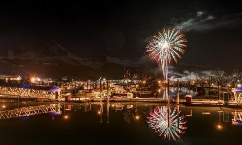 Colorful fireworks burst over a marina at night, reflecting in the water with boats and mountains in the background.