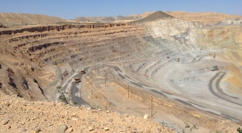 A vast open-pit mine with layered rock formations and heavy machinery working on the road. Clear blue sky above.