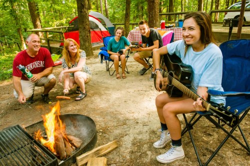 A group of friends enjoys a campsite, with one playing guitar by a fire while others relax and chat nearby.