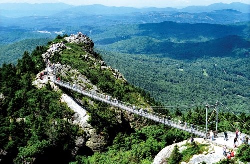 A suspension bridge connects a rocky peak to a forested mountain, with visitors enjoying the scenic view.