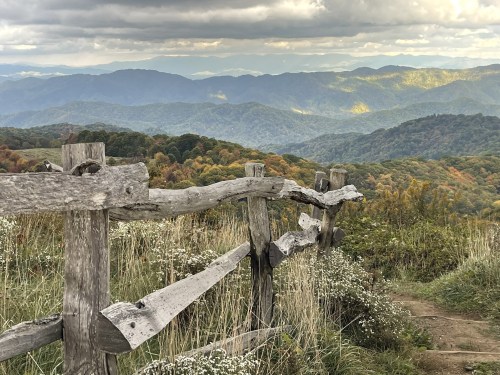 A wooden fence overlooks a scenic mountain landscape with rolling hills and colorful autumn foliage under a cloudy sky.