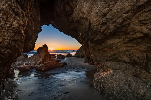 Sunset view through a rocky archway at the beach, with waves gently lapping at the shore.