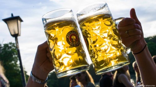 Two large beer mugs clink together, filled with golden beer, against a lively outdoor background.