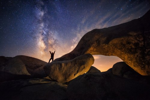 A silhouette of a person standing on rocks under a starry sky, with the Milky Way visible in the background.