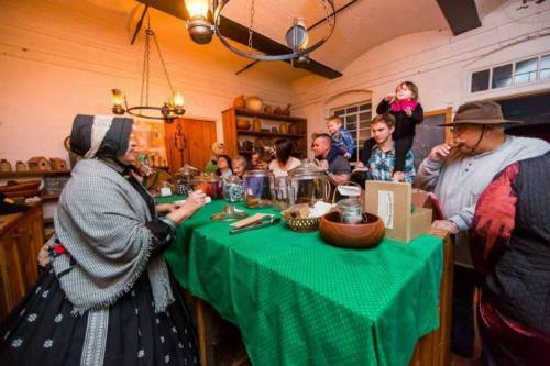 A historical kitchen scene with a costumed woman serving a group of people gathered around a green table.