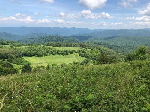 A panoramic view of rolling green hills and mountains under a blue sky with fluffy white clouds.