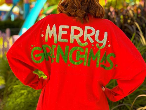 A person wearing a red shirt with the text "MERRY GRINCIMAS" in colorful letters, standing outdoors.