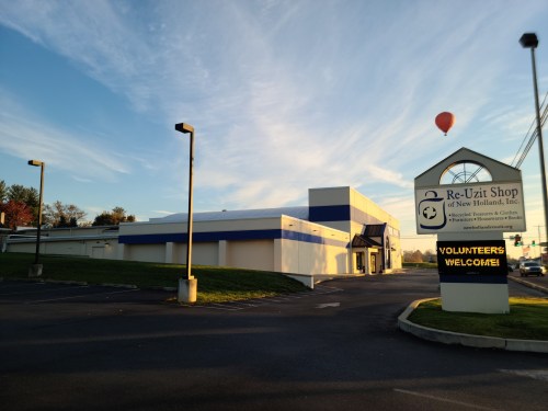 A building with a blue and white exterior, a sign reading "Volunteers Welcome," and a hot air balloon in the sky.
