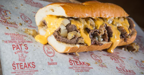 A loaded cheesesteak sandwich with melted cheese and onions on a hoagie roll, served on branded paper.