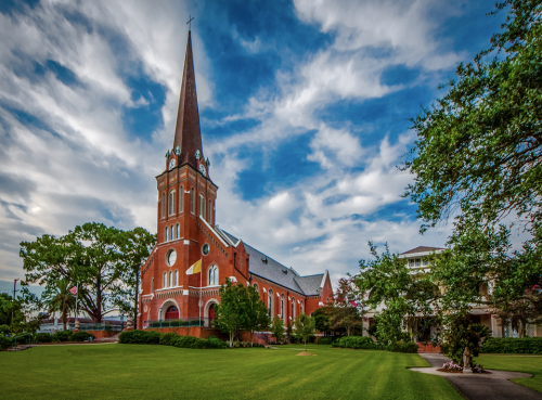 A red brick church with a tall spire, surrounded by green grass and trees under a blue sky with clouds.
