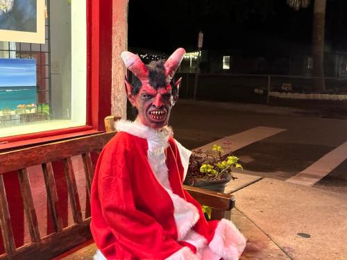 A figure in a red Santa suit with a demonic mask sits on a bench at night, surrounded by a quiet street.