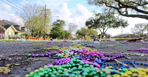 Colorful Mardi Gras beads scattered on a street, with trees and houses in the background under a blue sky.