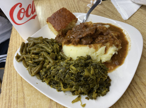 A plate of mashed potatoes with gravy, green beans, collard greens, and cornbread, alongside a cup of Coca-Cola.
