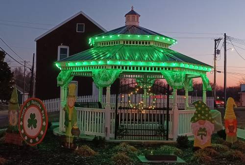 A gazebo decorated with green lights and St. Patrick's Day decorations at dusk, surrounded by festive gnomes and a sign.
