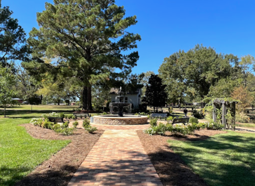 A sunny park scene featuring a brick path, fountain, benches, and lush greenery under a clear blue sky.