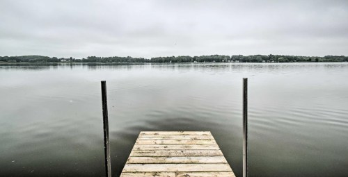 A wooden dock extends into a calm, gray lake surrounded by trees under a cloudy sky.