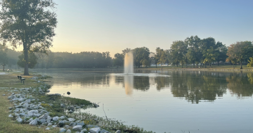 A serene park scene at dawn, featuring a calm lake with a fountain and trees reflecting in the water.