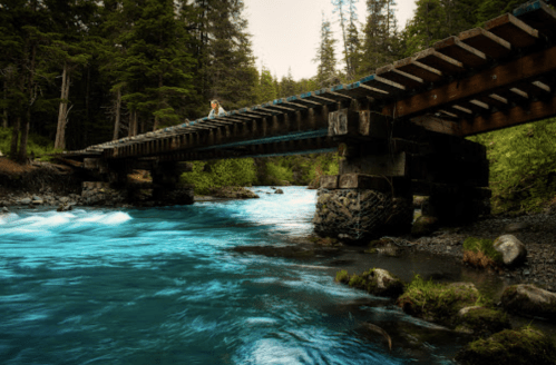 A person stands on a wooden bridge over a turquoise river, surrounded by lush green trees and rocky banks.