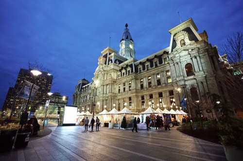 Historic building illuminated at dusk, with festive stalls and people walking in a lively urban square.