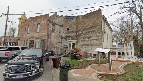A weathered brick building with a sloped roof, parked vehicles, and trash bins in a residential area.