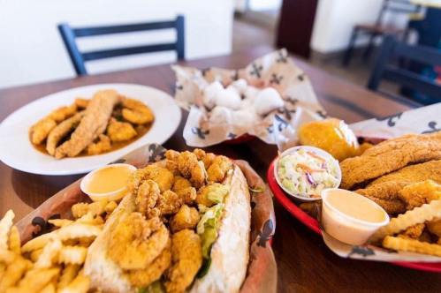A table with fried seafood, including shrimp po'boy, fried fish, fries, and hushpuppies, served with dipping sauces.