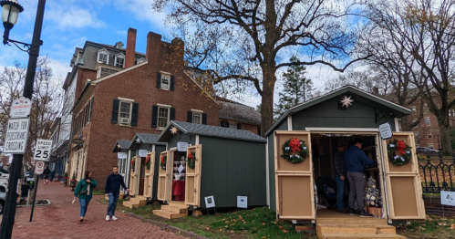A festive street scene with holiday market stalls, decorated with wreaths, near a historic brick building.