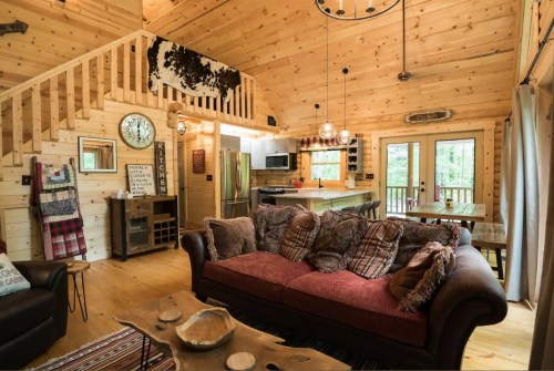 Cozy wooden cabin interior with a sofa, rustic decor, and a kitchen area, surrounded by large windows.