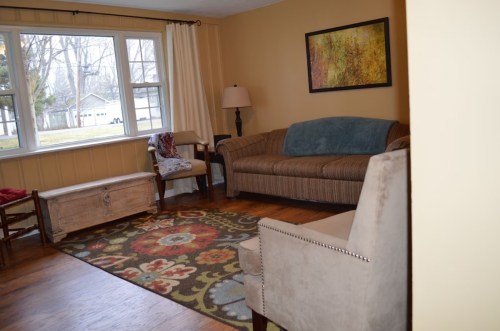 Cozy living room with a sofa, armchair, decorative rug, and a window overlooking a yard.