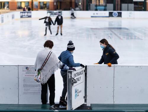 Two people in winter attire talk to a staff member at an ice skating rink, with skaters in the background.