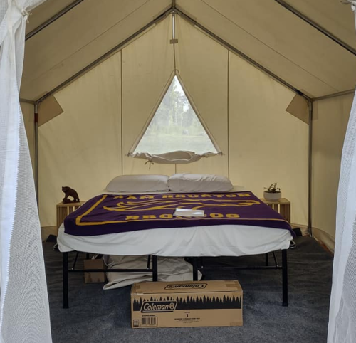 Interior of a tent featuring a bed with a purple blanket, side tables, and a box on the floor. Natural light enters through a window.