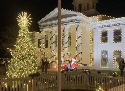 A festive scene featuring a decorated Christmas tree and Santa in front of the Swain County Heritage Museum, illuminated at night.