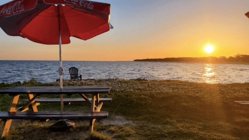 A picnic table with a red umbrella overlooks a sunset by the water, with a lone chair in the background.