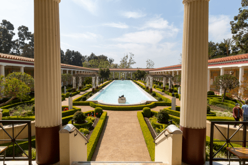 A serene garden with a central pool, surrounded by neatly trimmed hedges and classical columns under a clear blue sky.