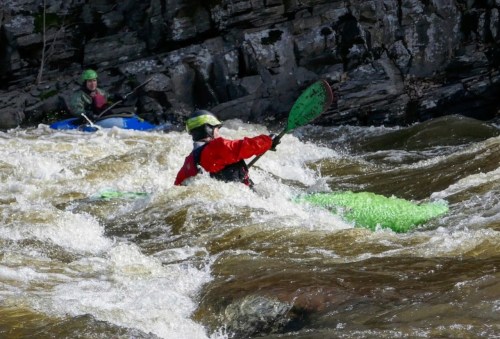 Two kayakers navigate turbulent waters, one in a green kayak and the other in blue, surrounded by rocky terrain.