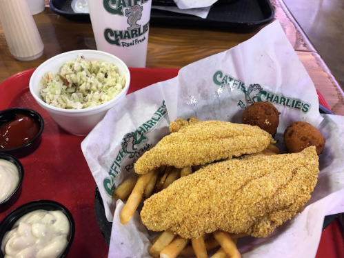 A tray with two fried catfish fillets, fries, coleslaw, hushpuppies, and dipping sauces on a red tray.