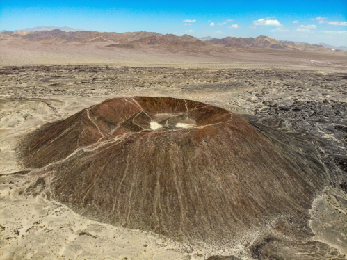 Aerial view of a large volcanic crater surrounded by arid landscape and distant mountains under a clear blue sky.