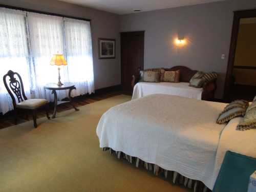 Cozy bedroom with two beds, a chair, a lamp, and large windows with lace curtains. Warm, inviting atmosphere.