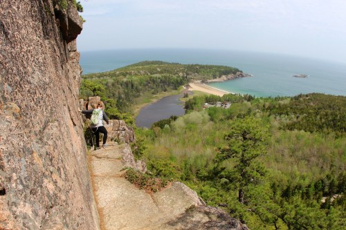 A person climbs a rocky cliff overlooking a lush green landscape and a beach along the coast.